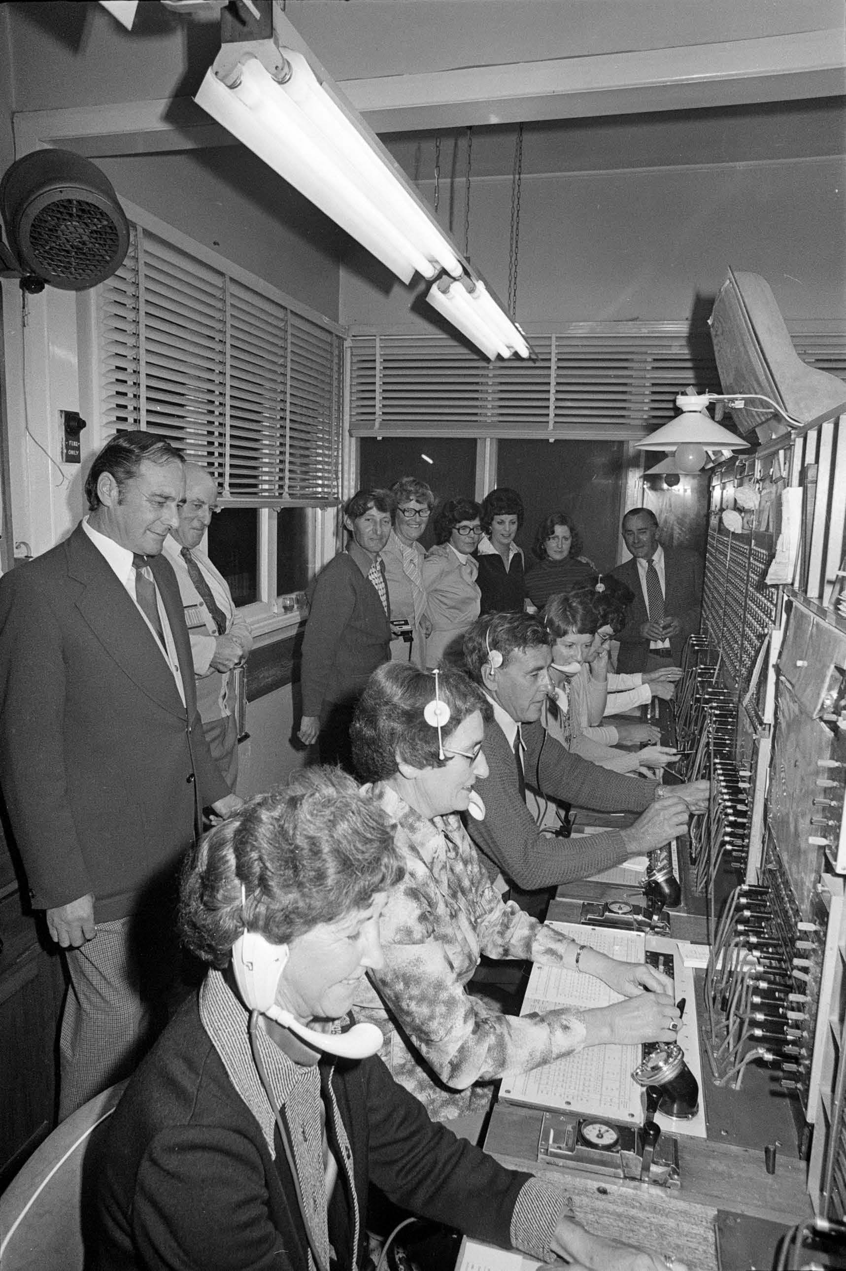 Black and white photo of a group of people in a telephone exchange, with many switchboards and operators.